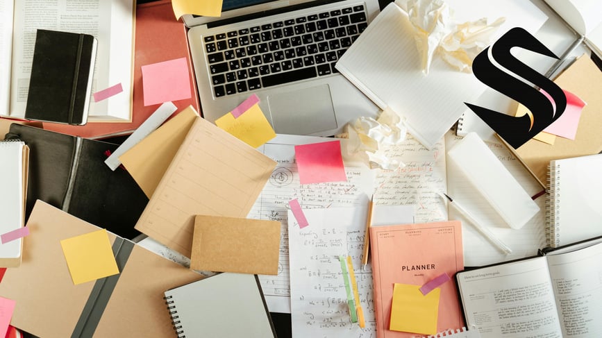 Cluttered desk with laptop, notes, and paperwork illustrating a CRM system overwhelmed by competing inputs and lack of ownership