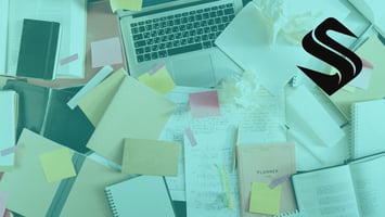 Cluttered desk with laptop, notes, and paperwork illustrating a CRM system overwhelmed by competing inputs and lack of ownership