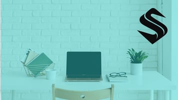 Minimal white desk with a laptop, notebook, glasses, and coffee cup arranged neatly against a white brick wall, representing an organized workspace.